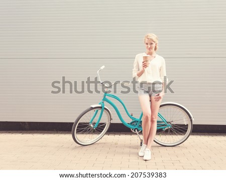 Young sexy blond woman standing near a green vintage bicycle and holding a cup of coffee have fun and good mood looking in camera and smiling
