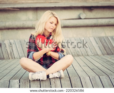 Sad lonely girl sitting on wood planks and hugging a big red heart