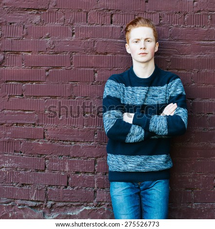 Young redhead man in a sweater and jeans standing next to a brick wall with folded arms