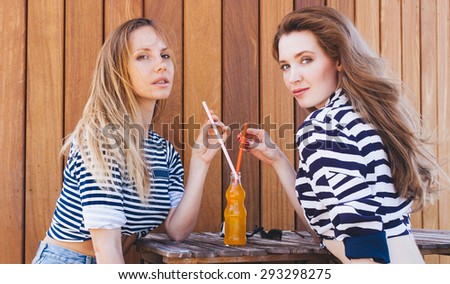 Two fashion beautiful girls sitting in a summer cafe and drink orange drink through a straw from a bottle. Sunny warm summer day. Outdoor.