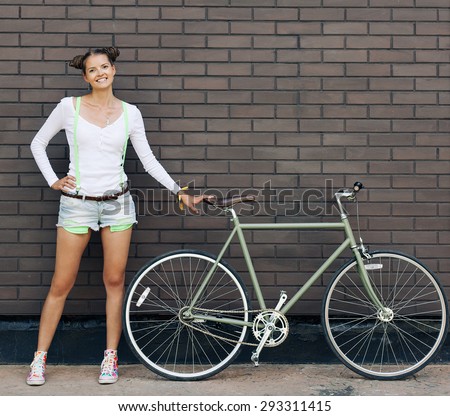 Pretty girl in shorts and t-shirt stands with bicycle fix gear near the brick wall of bright sunny day