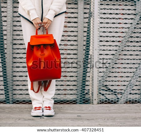 Fashionable beautiful big red backpack on the arm of the girl in a fashionable white sports suit, posing near the iron grid, wall on a warm summer night. Part of the body. Warm color. Close up.