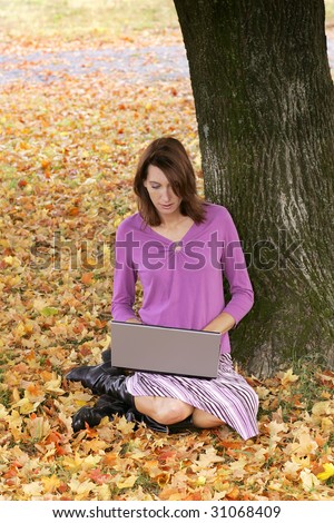 one young brunette woman sitting in the colorful fall leaves working on a laptop computer outdoors