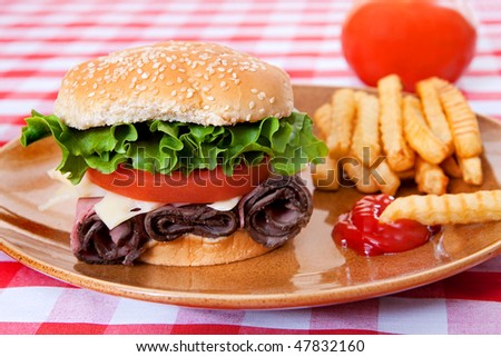 one large roast beef sandwich in sesame roll with tomatos and lettuce on plate with classic red and white checkered tablecloth
