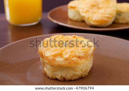 one yellow healthy broccoli and cheddar frittata muffin on a brown plate in front of a glass of orange juice and a stack of more muffins