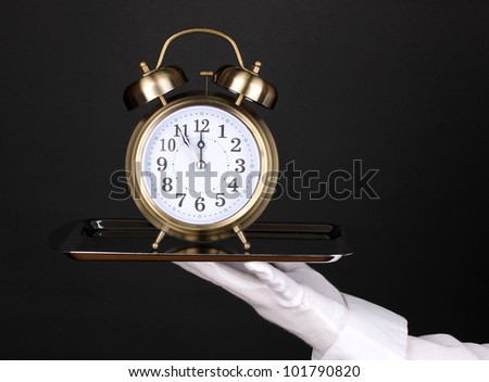 Hand in glove holding silver tray with alarm clock isolated on black