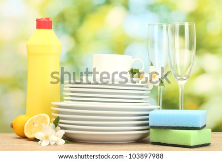 empty clean plates, glasses and cups with dishwashing liquid, sponges and lemon on wooden table on green background