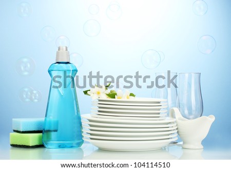 empty clean plates and glasses with dishwashing liquid, sponges and flowers on blue background