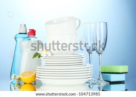 empty clean plates, glasses and cups with dishwashing liquid, sponges and lemon on blue background