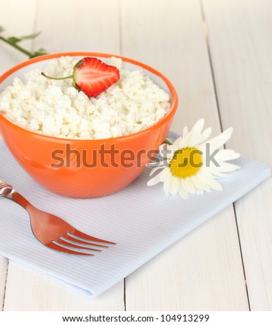 cottage cheese with strawberry in orange bowl, fork and flower on blue napkin on white wooden table close-up