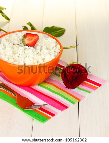 cottage cheese with strawberry in orange bowl, fork and flower on colorful napkin on white wooden table close-up