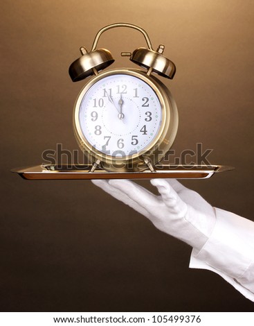 Hand in glove holding silver tray with alarm clock on brown background