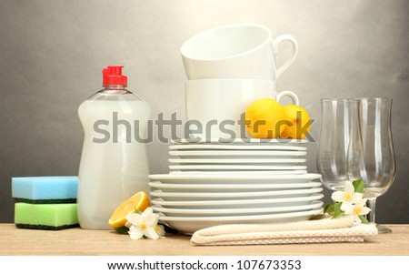 empty clean plates, glasses and cups with dishwashing liquid, sponges and lemon on wooden table on grey background