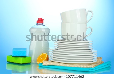 empty clean plates and cups with dishwashing liquid, sponges and lemon on blue background