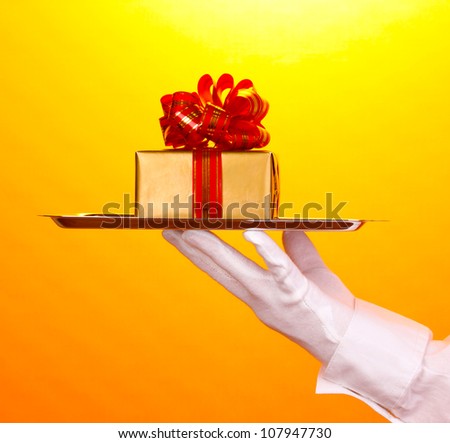 Hand in glove holding silver tray with giftbox on yellow background