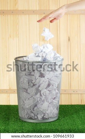 hand going garbage in metal trash bin from paper on grass on wooden background