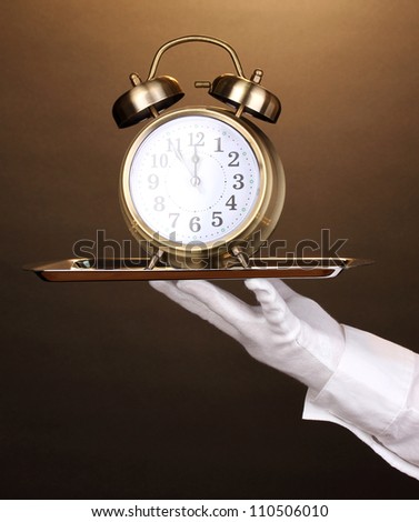 Hand in glove holding silver tray with alarm clock on brown background