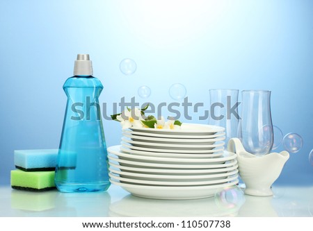empty clean plates and glasses with dishwashing liquid, sponges and flowers on blue background