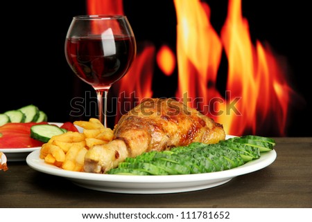 Roast chicken with french fries and cucumbers, glass of wine on wooden table  on fire background