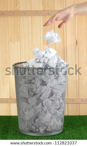 hand going garbage in metal trash bin from paper on grass on wooden background