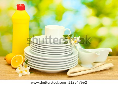 empty clean plates and cups with dishwashing liquid, flowers and lemon on wooden table on green background