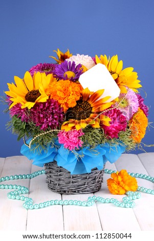 Beautiful bouquet of bright flowers with paper note on wooden table on blue background