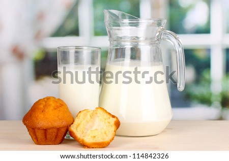 Pitcher and glass of milk with muffins on wooden table on window background