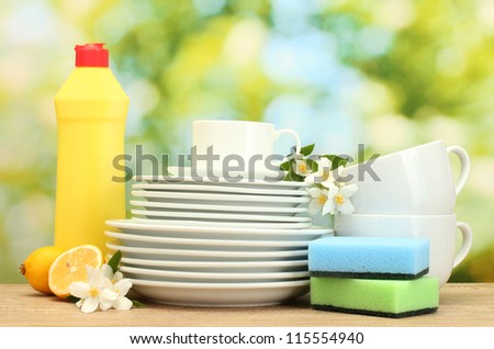 empty clean plates and cups with dishwashing liquid, sponges and lemon on wooden table on green background