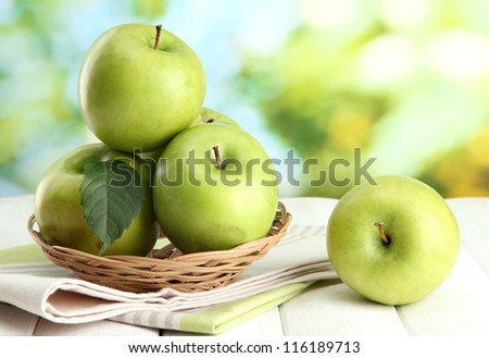 Ripe green apples with leaves in basket, on wooden table, on green background