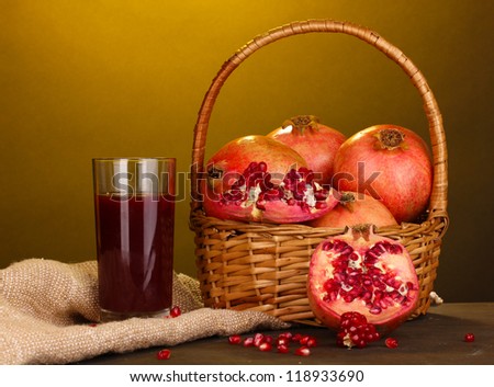 Ripe pomegranates on basket with glass of pomegranate juice on wooden table on yellow background