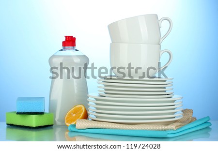 empty clean plates and cups with dishwashing liquid, sponges and lemon on blue background