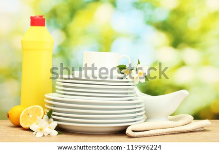 empty clean plates and cups with dishwashing liquid, flowers and lemon on wooden table on green background