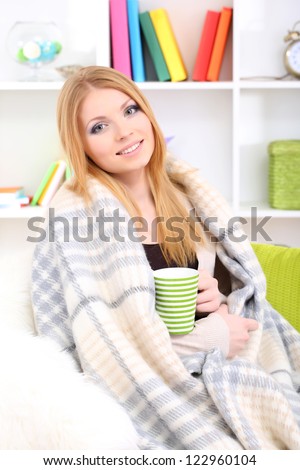 Attractive young woman sitting on sofa, holding cup with hot drink, on home interior background