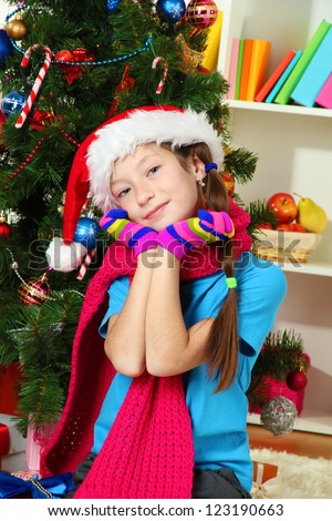 Little girl with pink scarf and multicolor gloves sitting near christmas tree