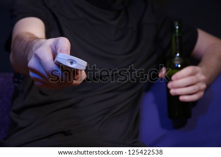 Man hand holding a TV remote control and beer bottle, on dark background