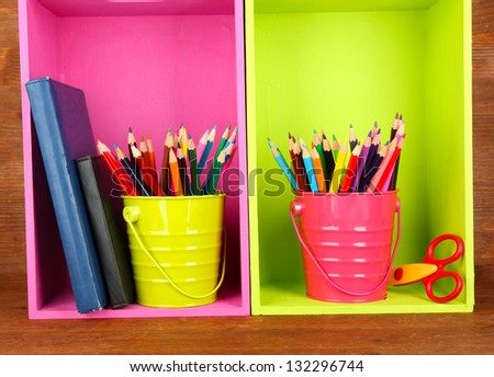Colorful pencils in pails on shelves with writing-pad on wooden background