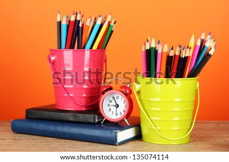 Colorful pencils in two pails with writing-pad on table on orange background