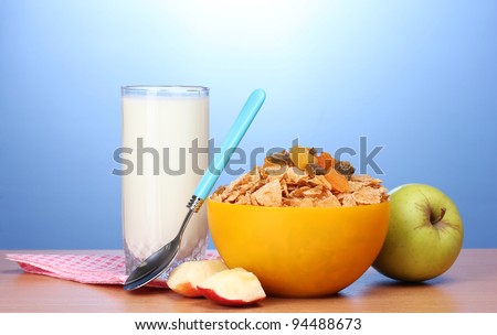 tasty cornflakes in yellow bowl, apples and glass of milk on wooden table on blue background