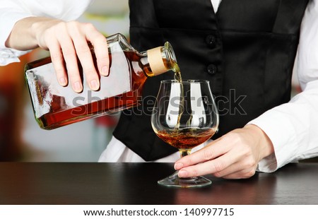 Barman hand with bottle of cognac  pouring drink into glass, on bright background