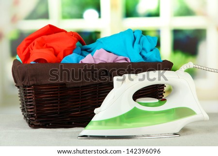 Steam iron and wicker basket with clothes, on bright background