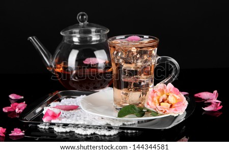 Kettle glass cup of ice tea from tea rose on metallic tray on napkin on black background