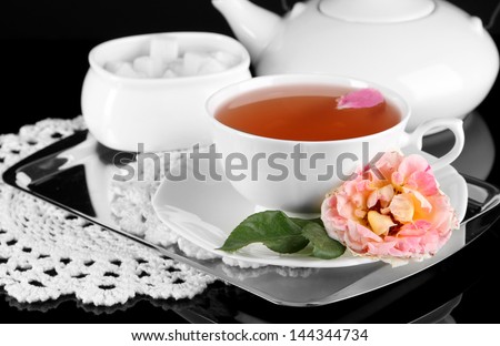 Kettle and cup of tea from tea rose on metallic tray on napkin black background