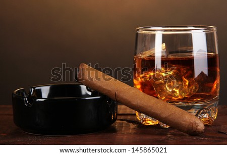 Brandy glass with ice and cigar on wooden table on brown background