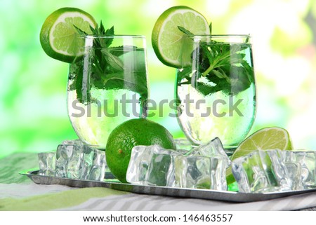 Glasses of cocktail with ice on metal tray on napkin on wooden table on nature background