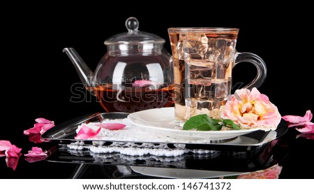 Kettle glass cup of ice tea from tea rose on metallic tray on napkin on black background