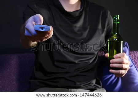 Man hand holding a TV remote control and beer bottle, on dark background