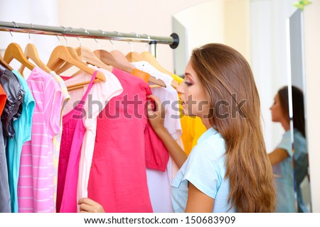 Beautiful girl chooses clothes on hangers on room background