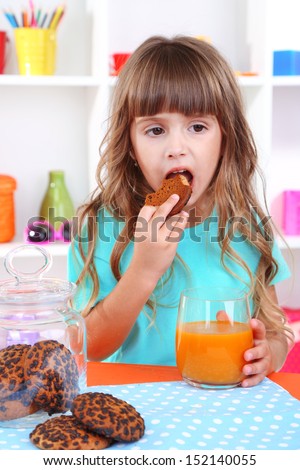 Little girl eating cookies and drinking juice sitting at table in room on shelves background