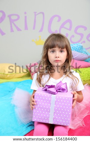 Little girl sitting on bed with gift in room on grey wall background