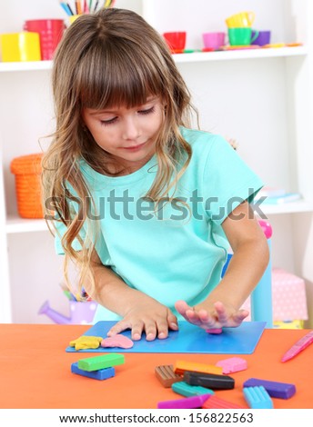 Little girl molds from plasticine sitting at table in room on shelves background